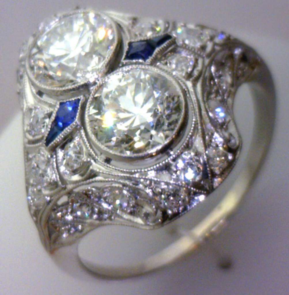 Close-up view of a platinum ring with two large round brilliant cut diamonds in full bezel setting surrounded by smaller diamonds and blue sapphires